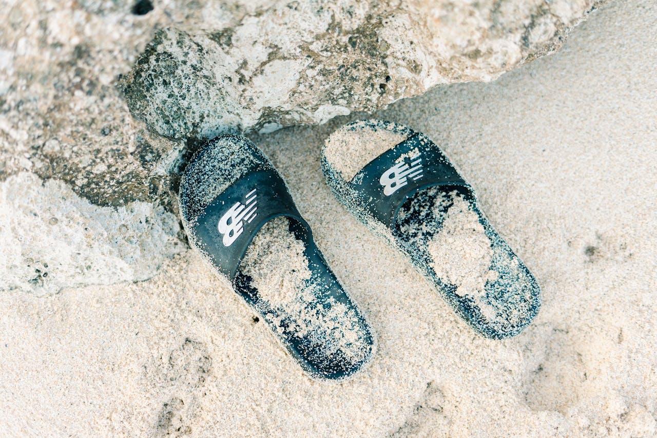 about-img-01 Two slippers covered in sand rest on a rocky beach shore captured in serene daylight.
